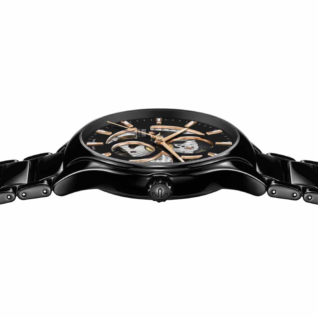 From a side angle, the RADO True Round Open Heart Automatic 40mm Watch is displayed, showcasing its black wristwatch design with a metallic band. The watch features a dark face accented with gold and high-tech ceramic construction, revealing visible gears that emphasize its intricate open heart design.