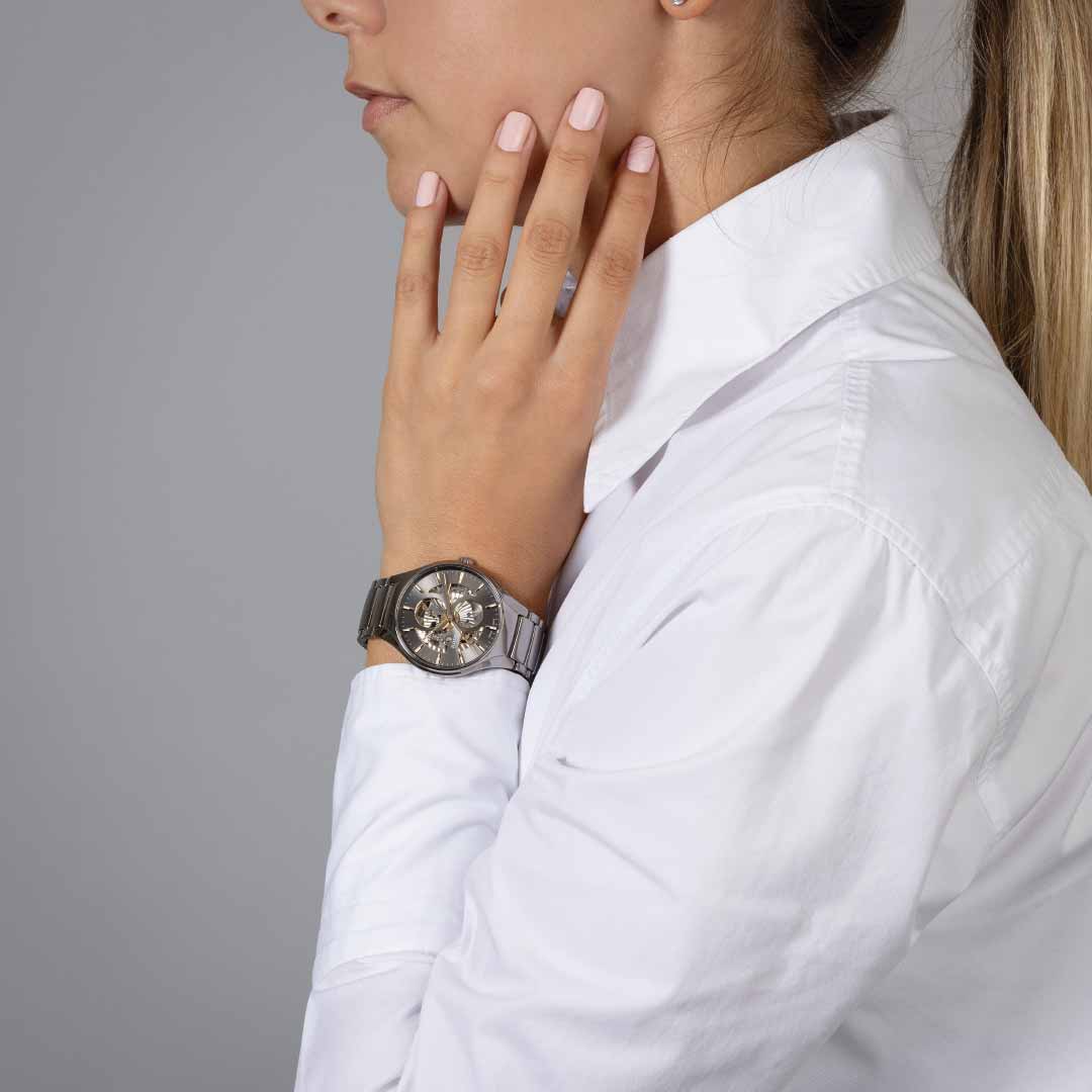 A person in a white shirt and wearing the RADO True Round Automatic Open Heart 40mm Watch rests their hand on their face. The watch, featuring a prominent face with multiple dials, pairs seamlessly with the person's long hair pulled back against the simple gray backdrop.