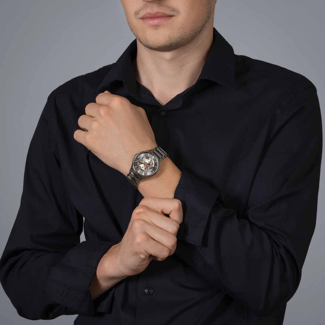 A person in a black shirt is adjusting their RADO True Round Automatic Open Heart 40mm Watch, showcasing the automatic Rado caliber R734 movement, on their left wrist against a plain gray background.