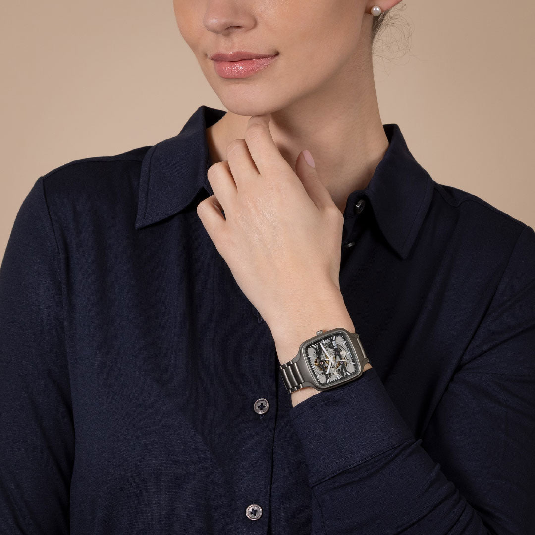 A woman in a dark navy button-up shirt touches her neck, highlighting the bold RADO True Square Automatic Skeleton 38mm Watch on her wrist. The striking silver watch by RADO stands out against the beige background.