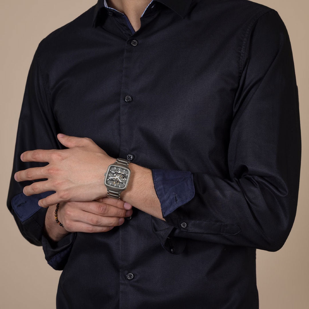 A person in a dark button-up shirt showcases the RADO True Square Automatic Skeleton 38mm Watch on their left wrist, with their right hand gently resting on their left arm against a neutral background.