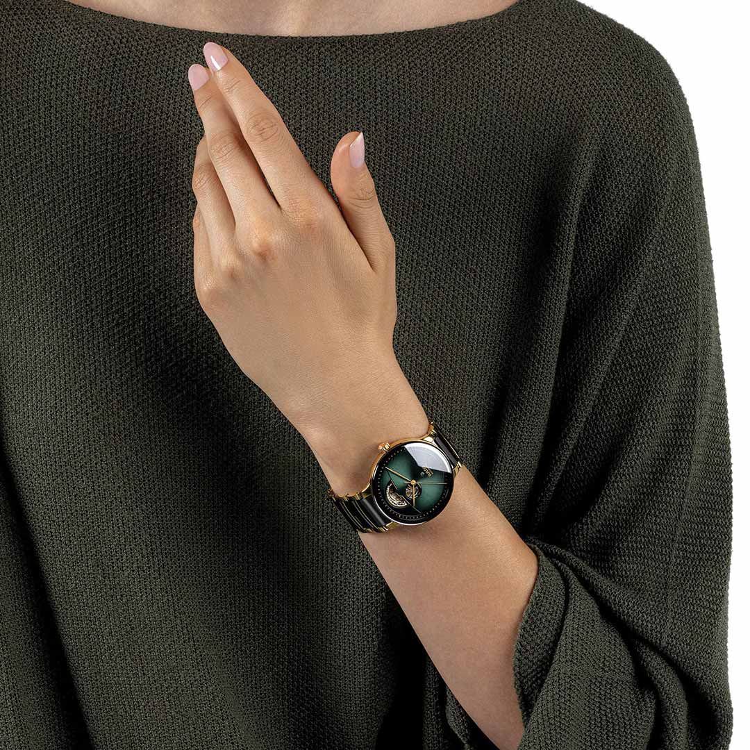 A person in an olive green sweater displays a RADO Centrix Automatic Open Heart 39.5mm Watch, featuring a gold band and a dark green watch face embellished with a decorative bee. The watch's automatic open heart movement ensures exceptional style and reliability, as the hand is elegantly raised near their chest.