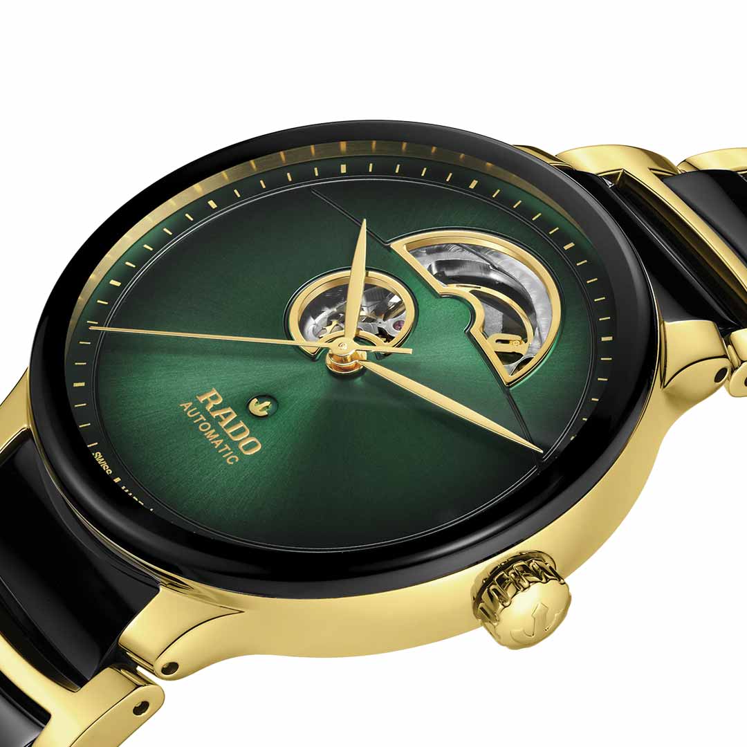 A detailed view of the RADO Centrix Automatic Open Heart 39.5mm Watch showcases its luxurious golden and black design. The timepiece features a captivating green dial that elegantly displays the inner workings, complemented by bold gold hour markers and the "Rado Automatic" label on the face.