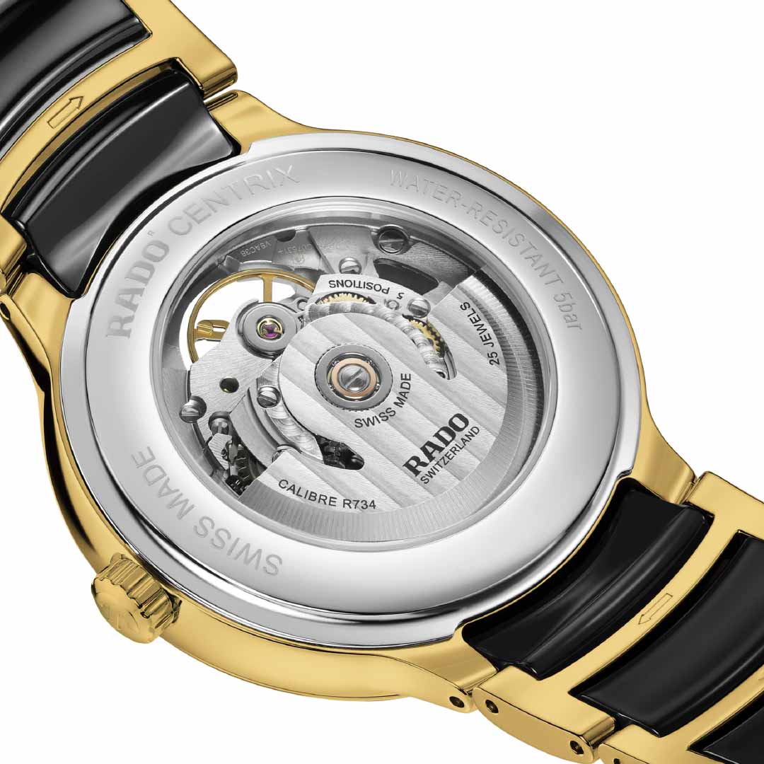 A close-up of the RADO Centrix Automatic Open Heart 39.5mm Watch showcases its intricate automatic movement. This luxurious timepiece features a gold and silver casing, complemented by a black ceramic band. The inscription "Swiss Made, 5 Bar" highlights its quality, while its impressive 80-hour power reserve caters to discerning watch enthusiasts.