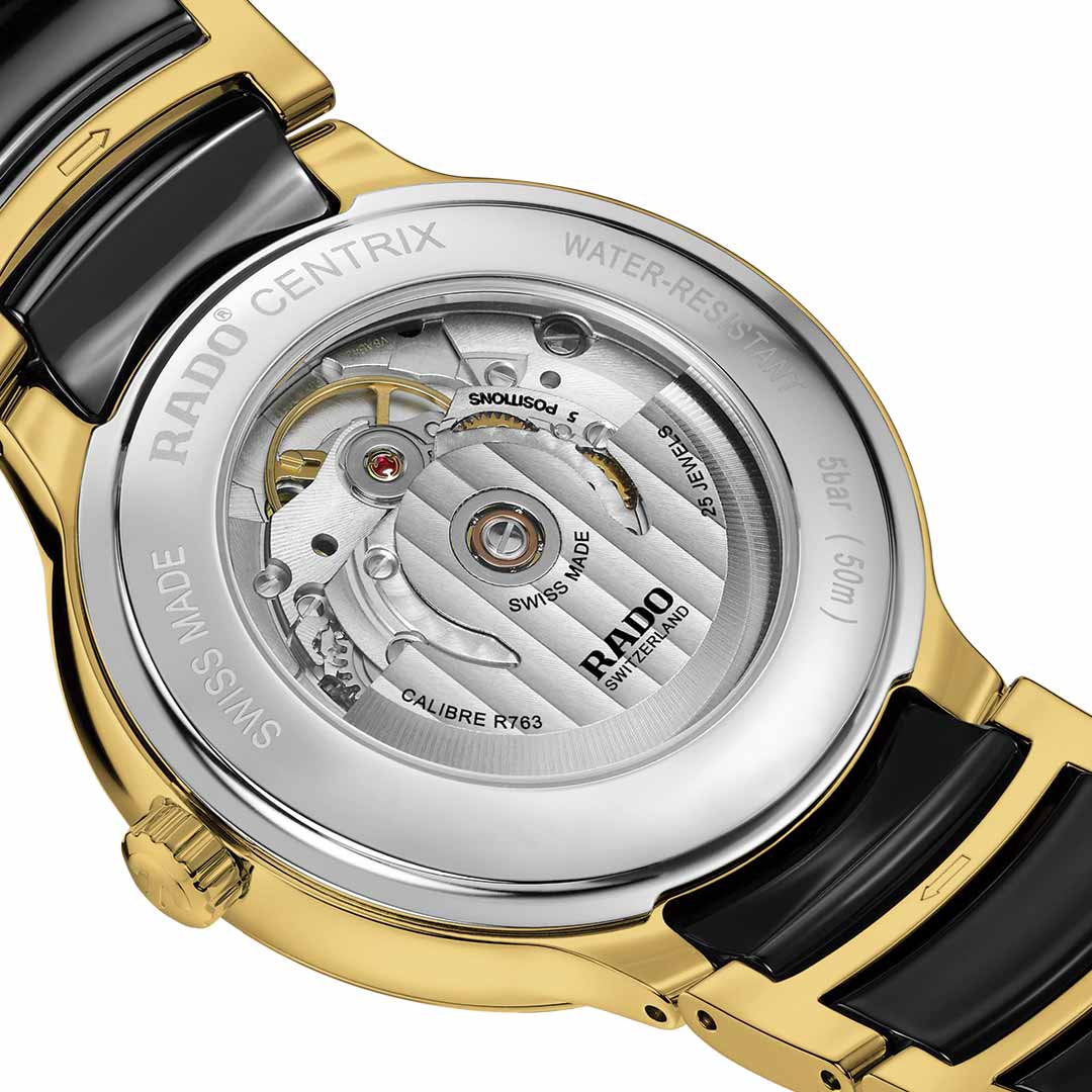 A detailed view of the RADO Centrix Diamonds Automatic 39.5mm watch back showcases the visible mechanical movement, adorned with "Swiss Made" and "5 bar (50m)" inscriptions. The timepiece features a yellow gold-coloured stainless steel case paired with a black ceramic bracelet, emphasizing the sophisticated craftsmanship of its antimagnetic Nivachron™ hairspring.