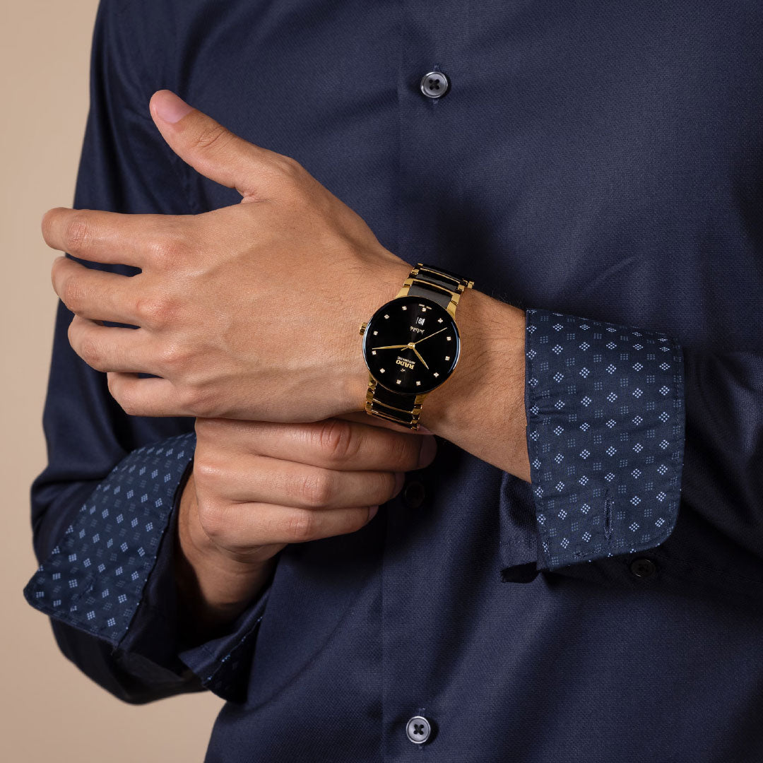 A person wearing a dark blue patterned dress shirt adjusts a RADO Centrix Automatic Diamonds 39.5mm watch, featuring black high-tech ceramic and yellow gold-coloured stainless steel, on their left wrist; only the hands and watch are visible.