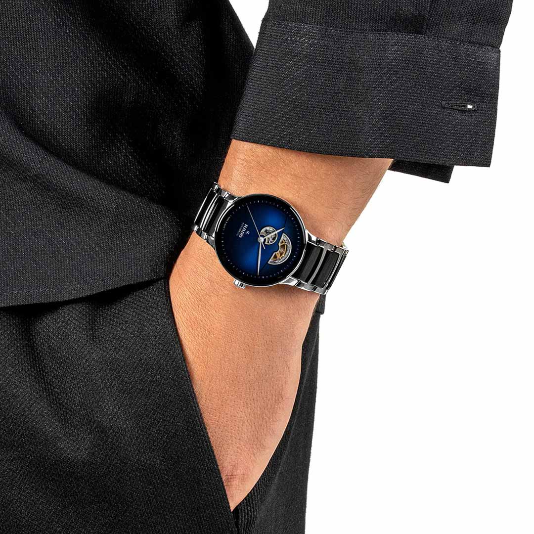 A person in a black suit effortlessly slips their hand into their pocket, displaying a RADO Centrix Open Heart Automatic 39.5mm watch with a silver finish and blue dial showcasing visible gears. The precision of this timepiece is enhanced by its Nivachron hairspring, while the 80-hour power reserve complements the refined texture of their outfit.