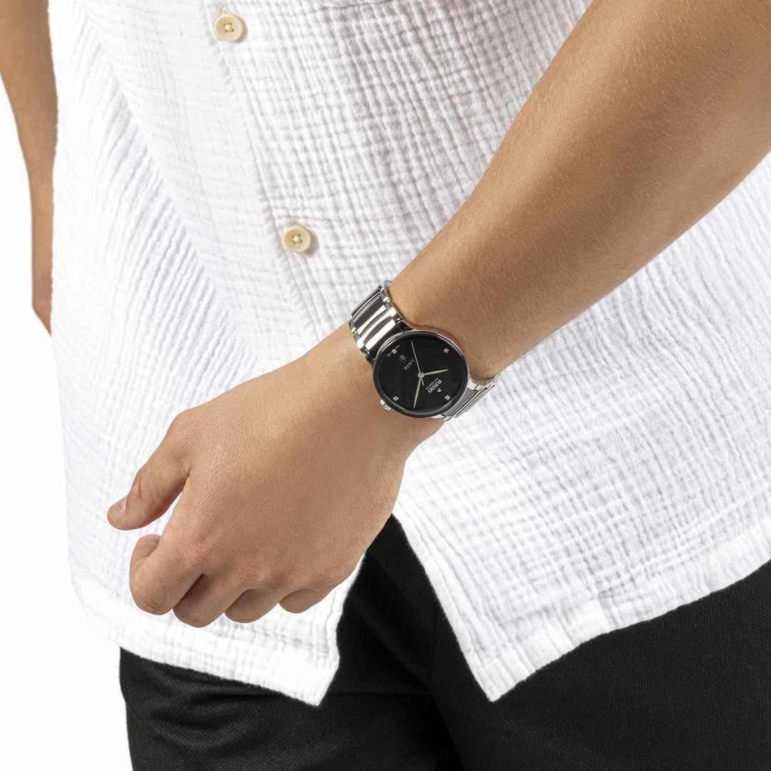 A person wearing a white textured shirt and dark pants is shown from the neck down. On their left wrist, they sport a silver RADO Centrix Diamonds Automatic 39.5mm watch with a black face. Their hand, relaxed by their side, showcases the elegance of high-tech ceramic craftsmanship.