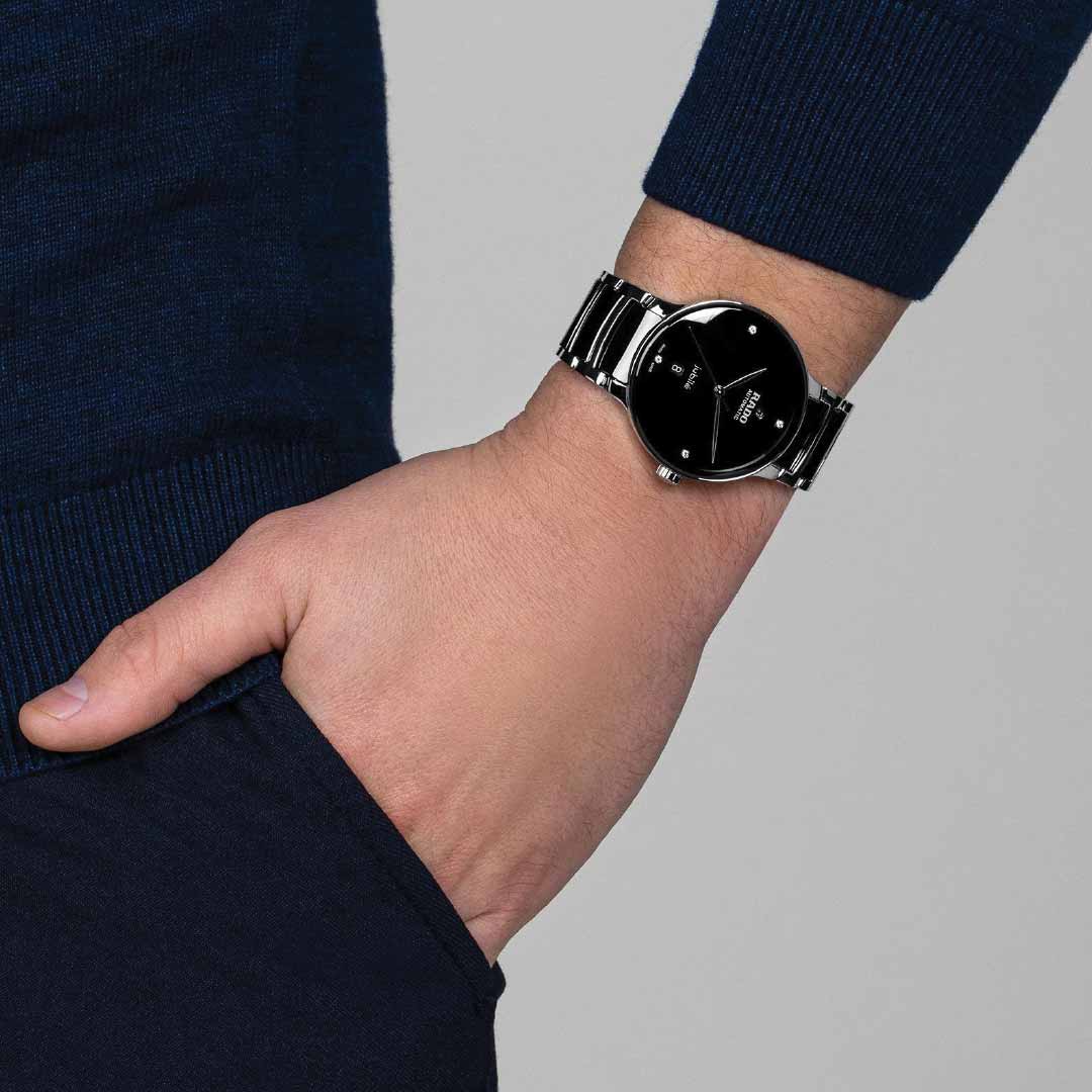 A person wearing a dark blue sweater has their hand in the pocket of dark pants. On their wrist is the sleek RADO Centrix Diamonds Automatic 39.5mm Watch, which showcases a shiny metal strap and Nivachron™ hairspring, effortlessly combining elegance with high-tech ceramic sophistication.