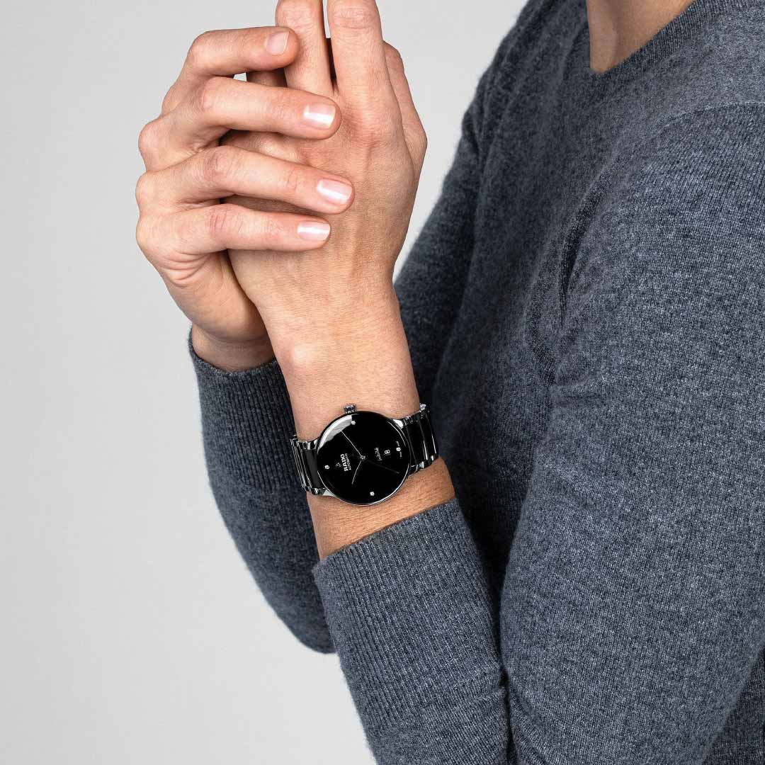 A person in a gray sweater holds a smartphone near their face with both hands. They showcase the sleek RADO Centrix Diamonds Automatic 39.5mm Watch, which features a high-tech ceramic band and an elegant black dial. The background is a plain light gray.