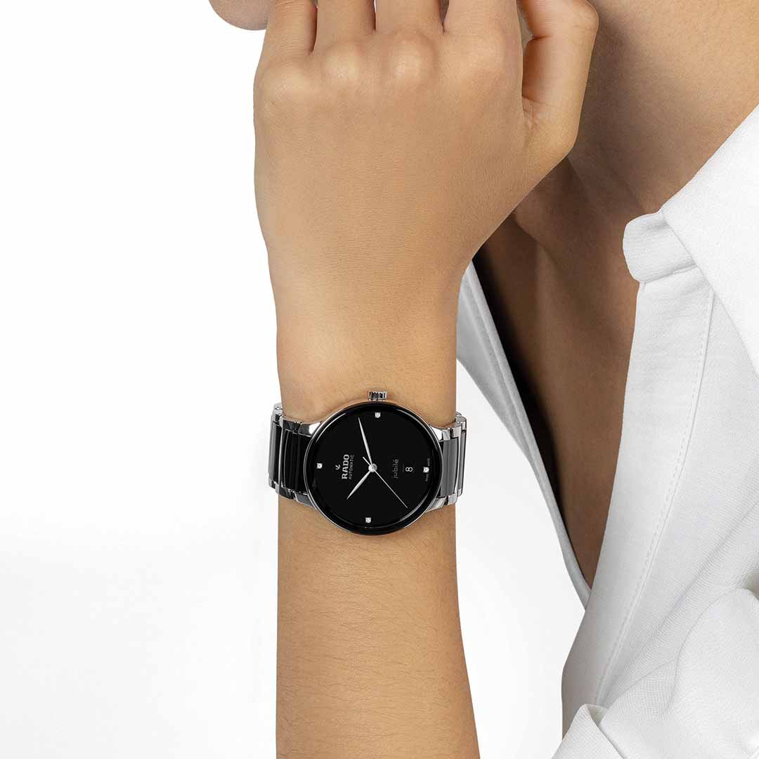 A person wearing a RADO Centrix Diamonds Automatic 39.5mm Watch rests their chin on their hand. The watch, showcasing a sleek black dial and featuring the innovative Nivachron™ hairspring, perfectly complements their white shirt with its minimalistic design.