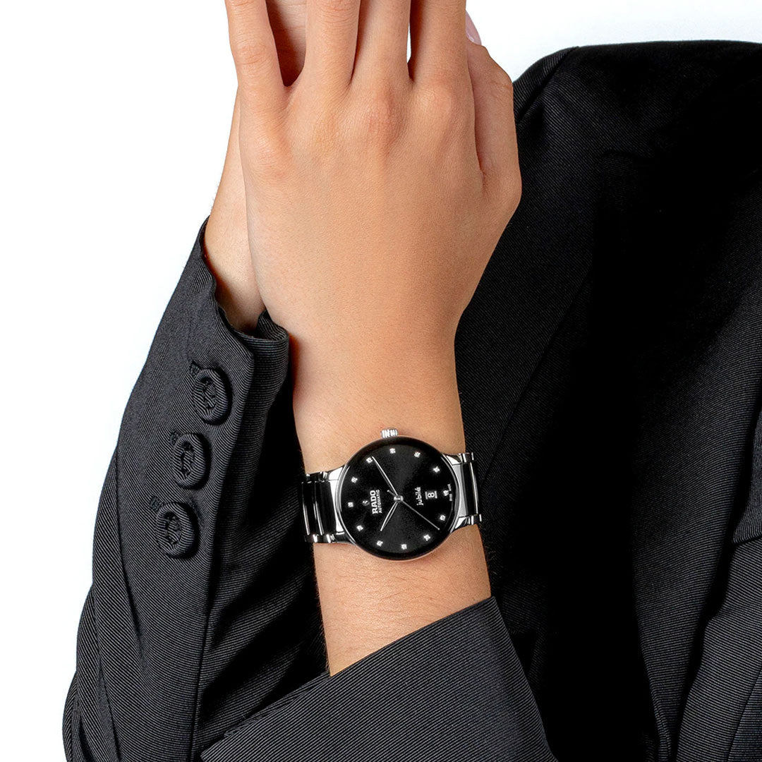 A person in a black suit showcases their wrist, wearing a RADO Centrix Automatic Diamonds 39.5mm watch with a black dial and metal band. Their hand is raised near their face, partially concealing it.