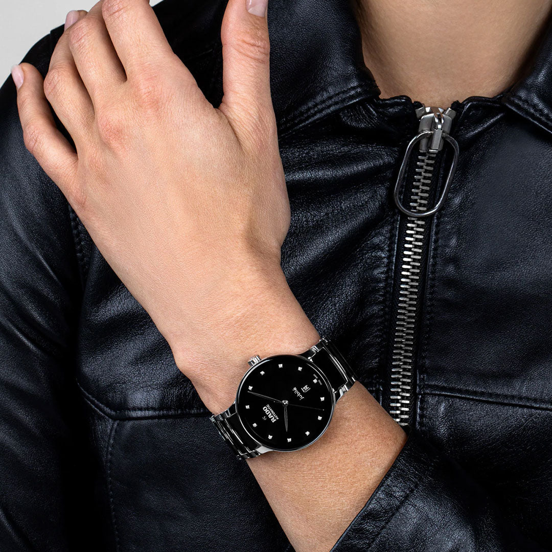 A person in a black leather jacket wears a RADO Centrix Automatic Diamonds 39.5mm watch—a sleek silver timepiece with diamond accents. Their hand rests on their chest, partially unzipping the jacket.