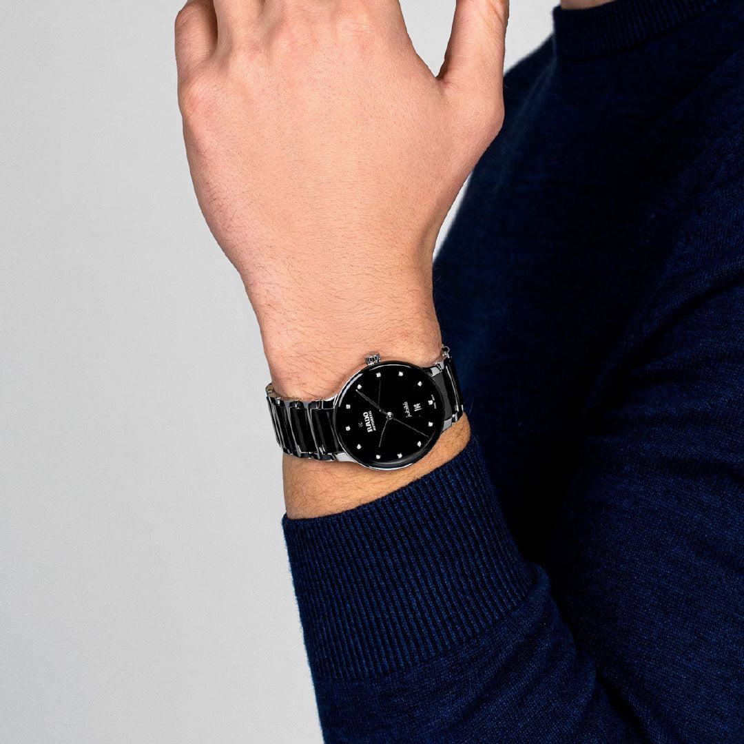 A person in a dark blue long-sleeve sweater displays their left wrist, showcasing the RADO Centrix Automatic Diamonds 39.5mm watch with a round face and metal band, against a plain light background.