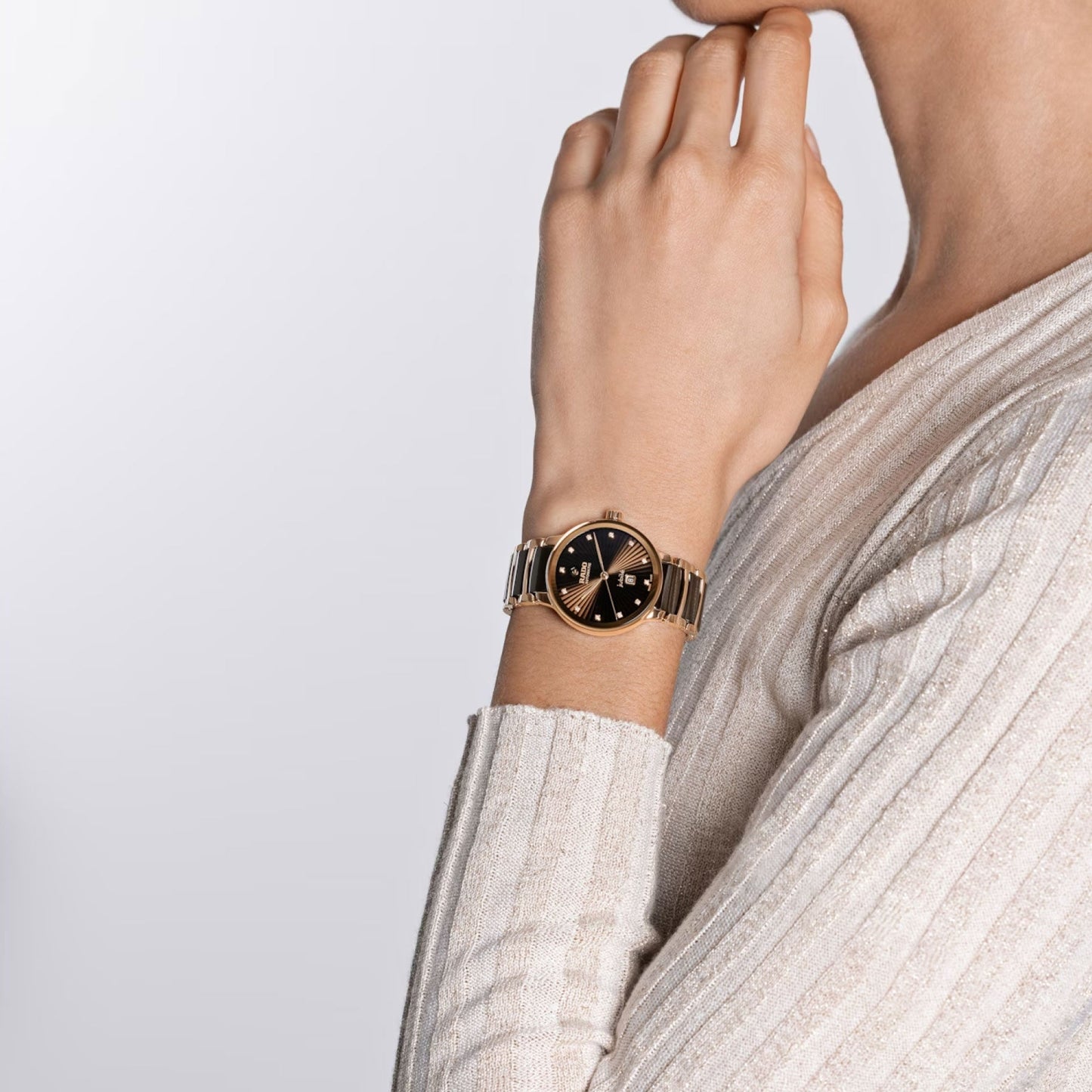 The image displays an individual from the shoulders up, dressed in a ribbed cream sweater. On their left wrist, raised to touch their chin, they are wearing a RADO Centrix Diamonds Automatic 30.5mm Watch with a black face. The background is a simple light gray.