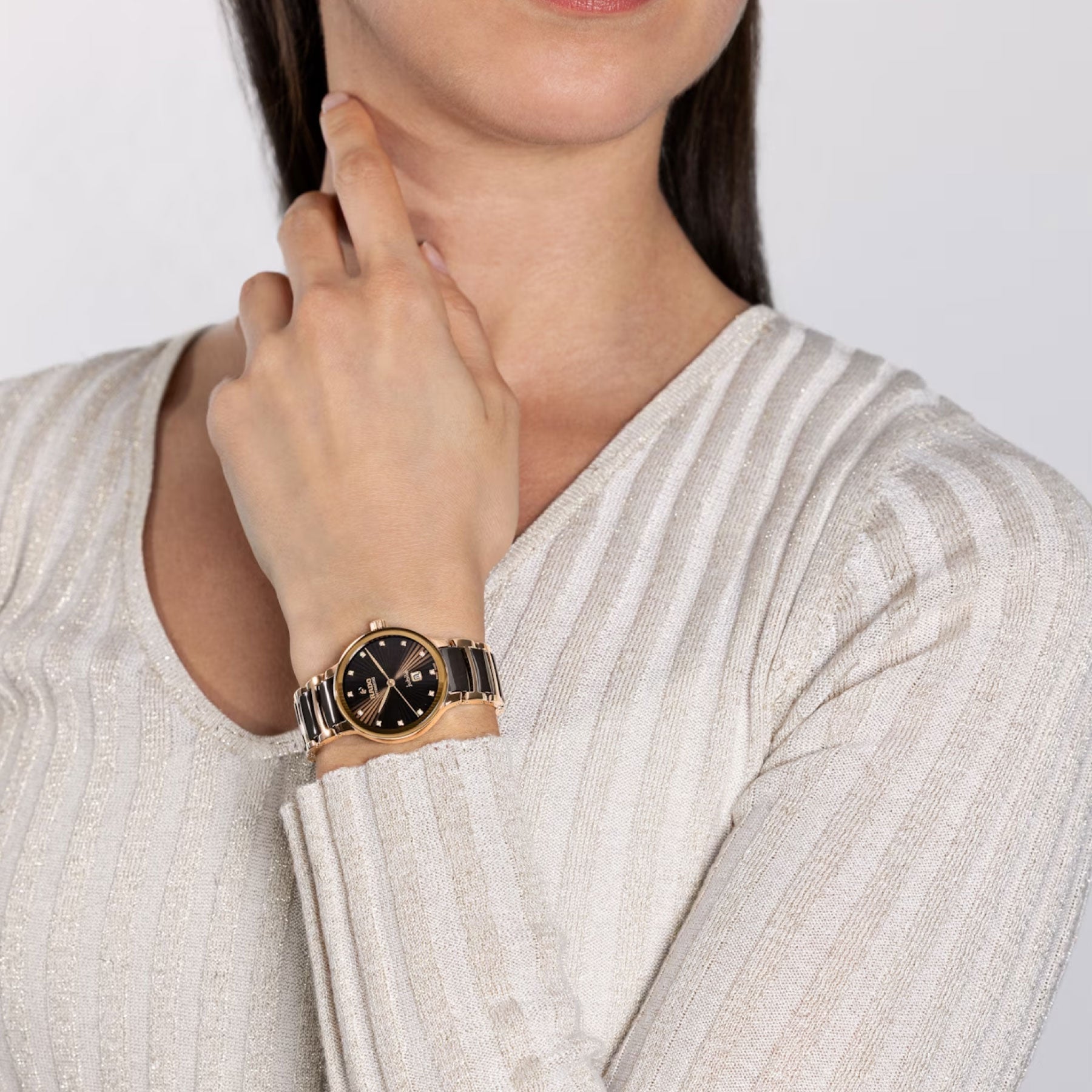 A person wearing a light-colored, textured top showcases the RADO Centrix Diamonds Automatic 30.5mm Watch, which features a striking black and gold face under sapphire crystal. The watch's two-tone metal band elegantly gleams with hints of rose-gold-colored PVD-coated stainless steel as they pose thoughtfully against a plain background.