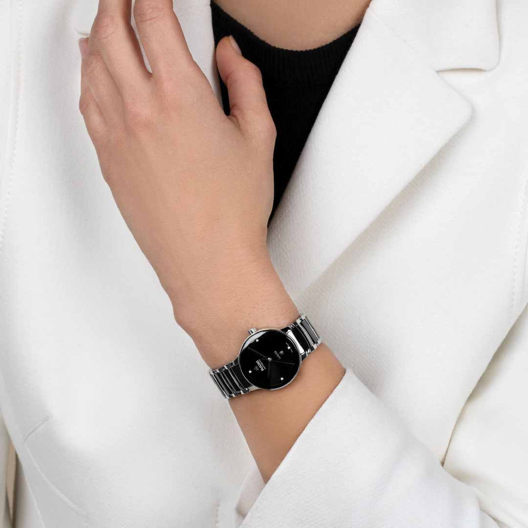 A person wearing a white blazer and a RADO Centrix Diamonds Automatic 30.5mm watch with a silver metal band featuring the antimagnetic Nivachron™ hairspring, their hand raised near their face against a plain background.