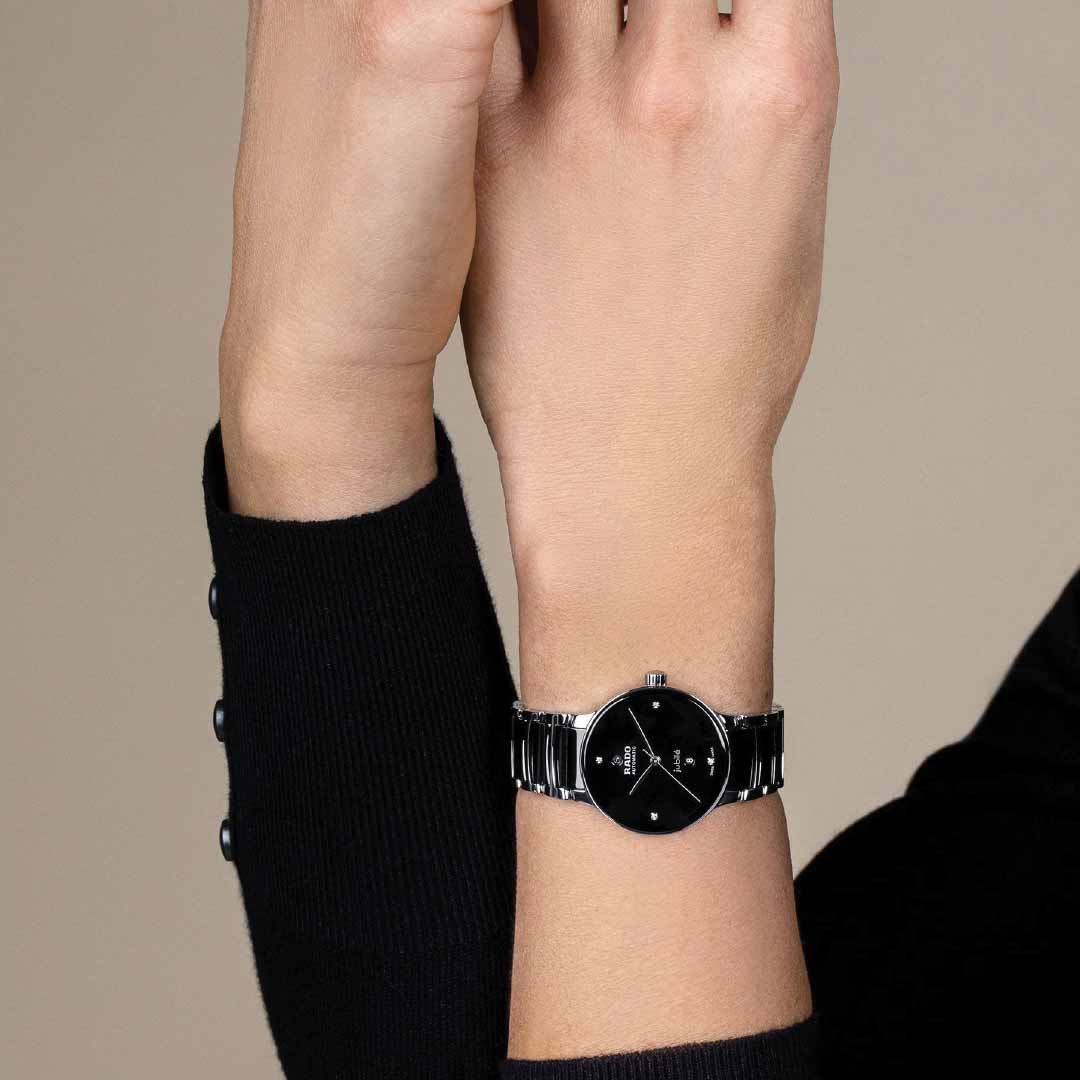 A person wearing a black sweater showcases a stylish RADO Centrix Diamonds Automatic 30.5mm watch, featuring a silver design with a black dial and an antimagnetic Nivachron™ hairspring for precision. The background is a plain, neutral color.