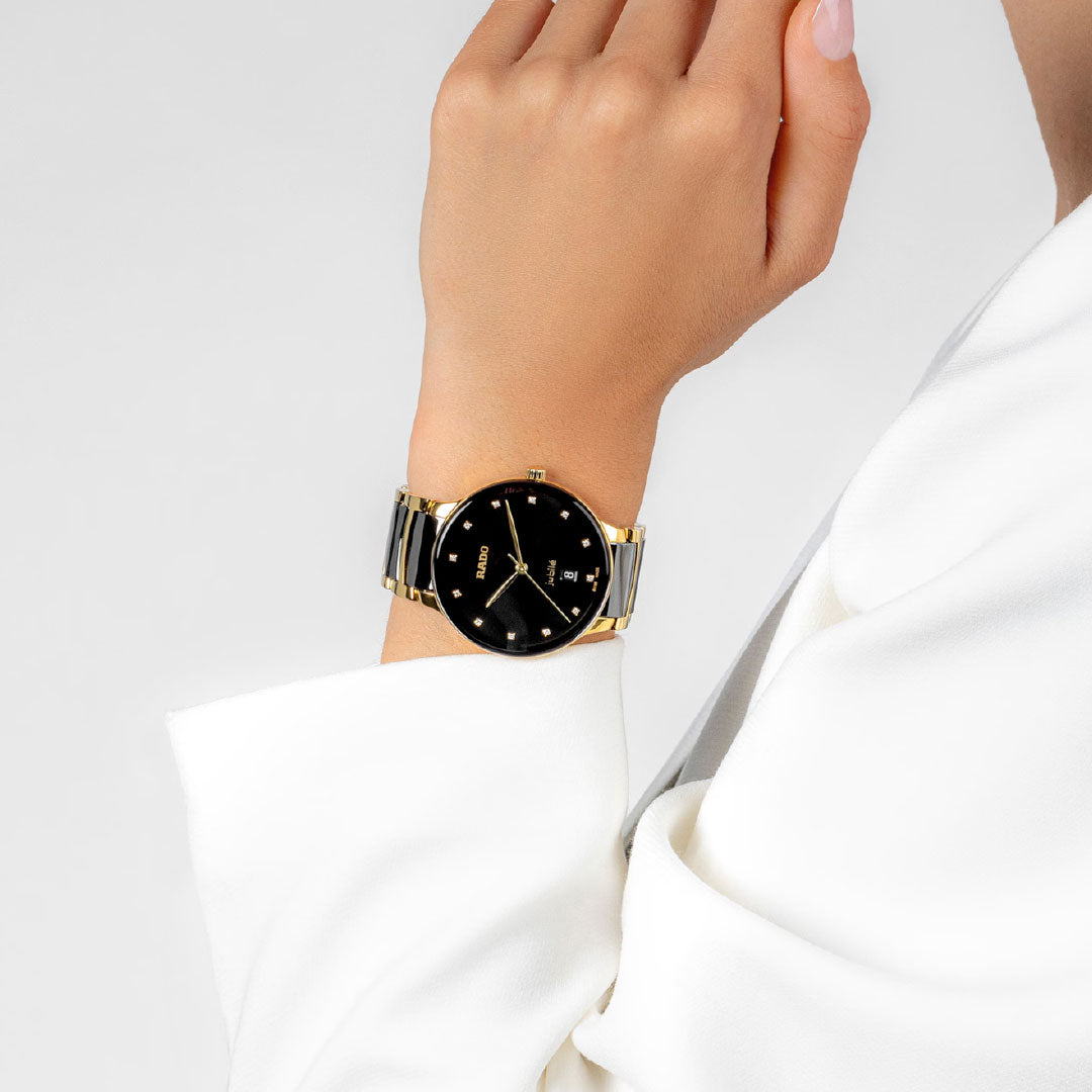 A person in a white blazer showcases the RADO Centrix Diamonds Quartz 39.5mm watch, featuring black high-tech ceramic and yellow gold-coloured PVD-coated stainless steel on the bracelet, worn on their left wrist against a plain light background.