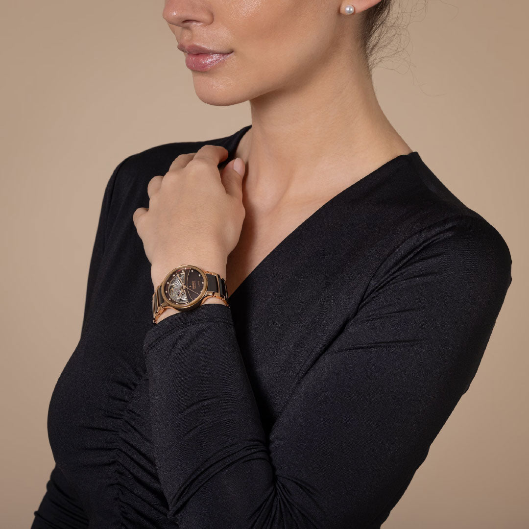 A woman in a black long-sleeve dress wears the RADO Centrix Automatic Diamonds Open Heart 35mm Watch and a pearl stud earring, posing with her hand near her collarbone against a beige background. Her face is partially visible.