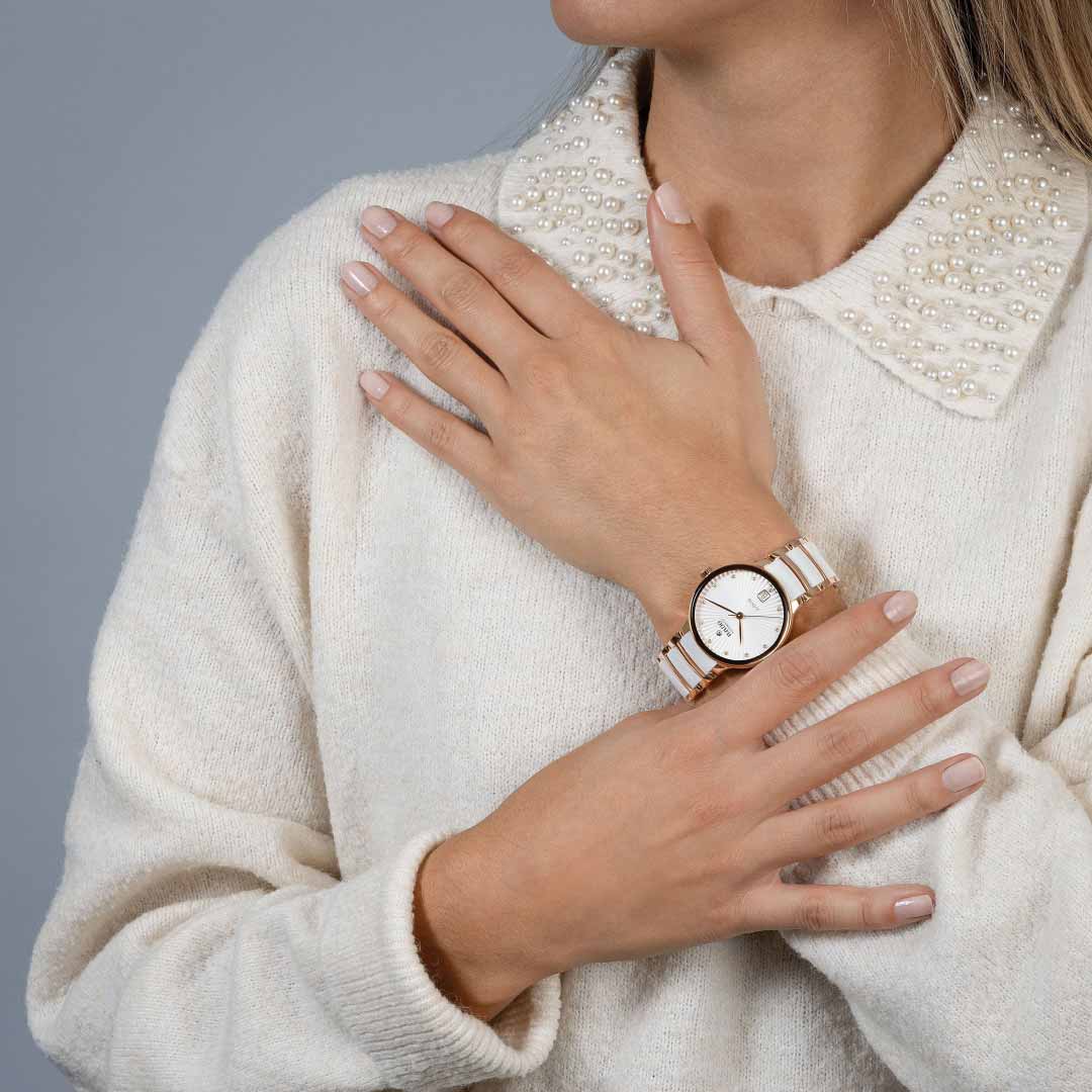 A person wearing a beige sweater with a pearl-studded collar showcases their RADO Centrix Diamonds Automatic 35mm Watch, which features a gold and silver band with a white face. The watch boasts sapphire crystal and rose-gold-coloured PVD-coated stainless steel, while their right hand gently rests on the left arm.