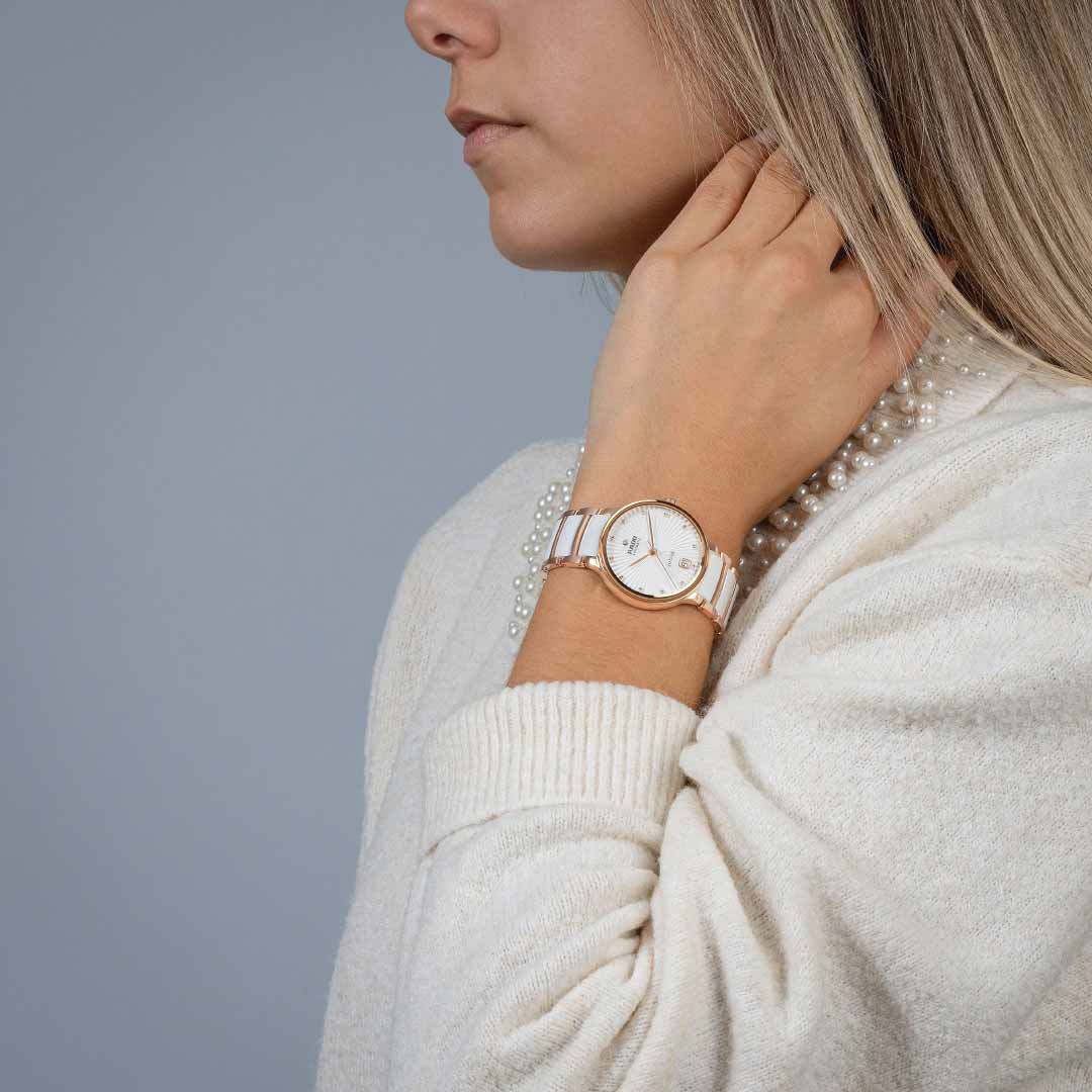 A person with shoulder-length blonde hair adjusts a pearl necklace, dressed in a white sweater. On their wrist is the RADO Centrix Diamonds Automatic 35mm Watch, featuring a rose-gold-coloured PVD-coated stainless steel case and a white strap, set against a plain grey background.