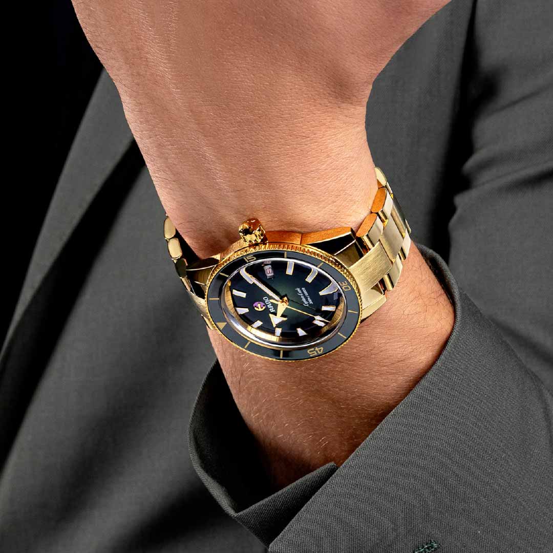 A close-up of a person's wrist showcases the RADO Captain Cook Automatic 42mm Watch with its sleek black dial. This gold timepiece from RADO features automatic movement and water resistance, beautifully complementing the metal band. The person is dressed in a gray suit jacket, enhancing the sophisticated look.