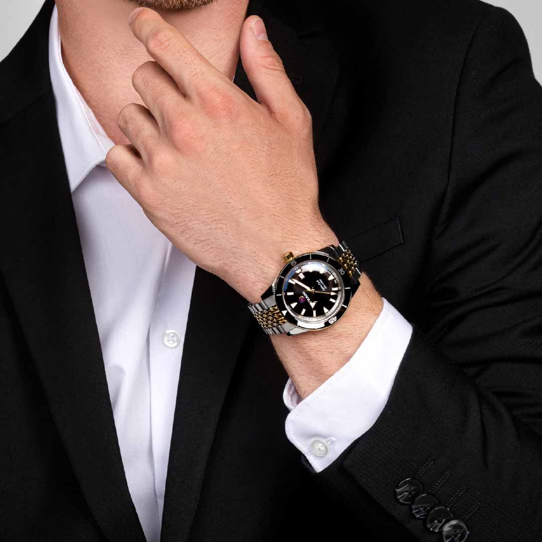 Dressed in a black suit and white shirt, the individual poses thoughtfully with a hand to their chin, highlighting the RADO Captain Cook Automatic 42mm Watch. This stylish timepiece features a water-resistant design and a black-and-gold metal band that exudes vintage charm.