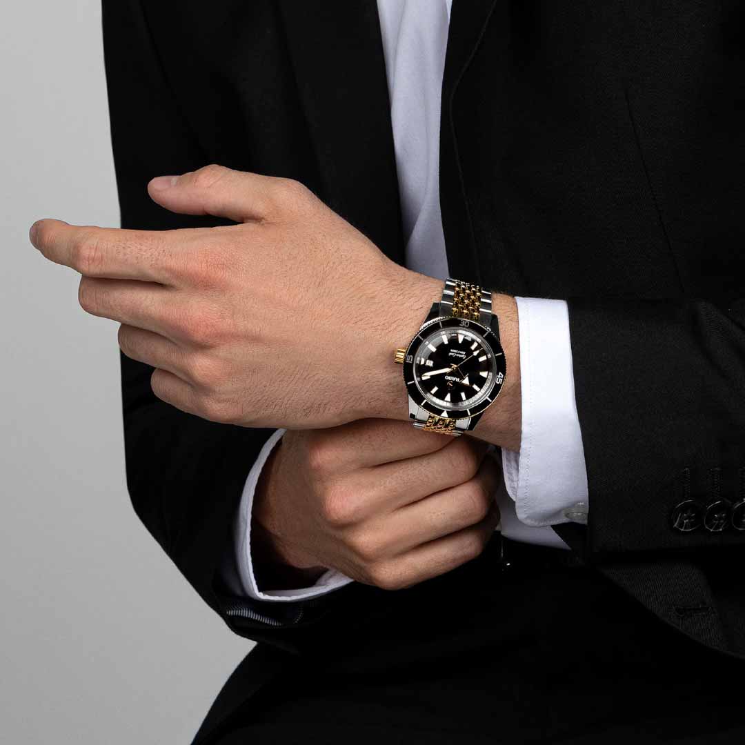 A man in a black suit with a white shirt adjusts his RADO Captain Cook Automatic 42mm watch. His left hand is near the timepiece on his right wrist, showcasing its gold and black elegance and water-resistant feature against a neutral background.