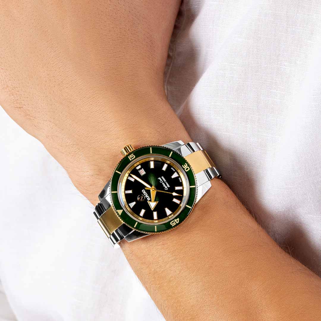 A wrist wearing a RADO Captain Cook Automatic 42mm Watch with a metallic strap. The lacquered green-to-black gradient dial stylishly features gold hour markers and a rotating bezel. The person's sleeve is rolled up, highlighting the watch and its EasyClip system for effortless strap changes.