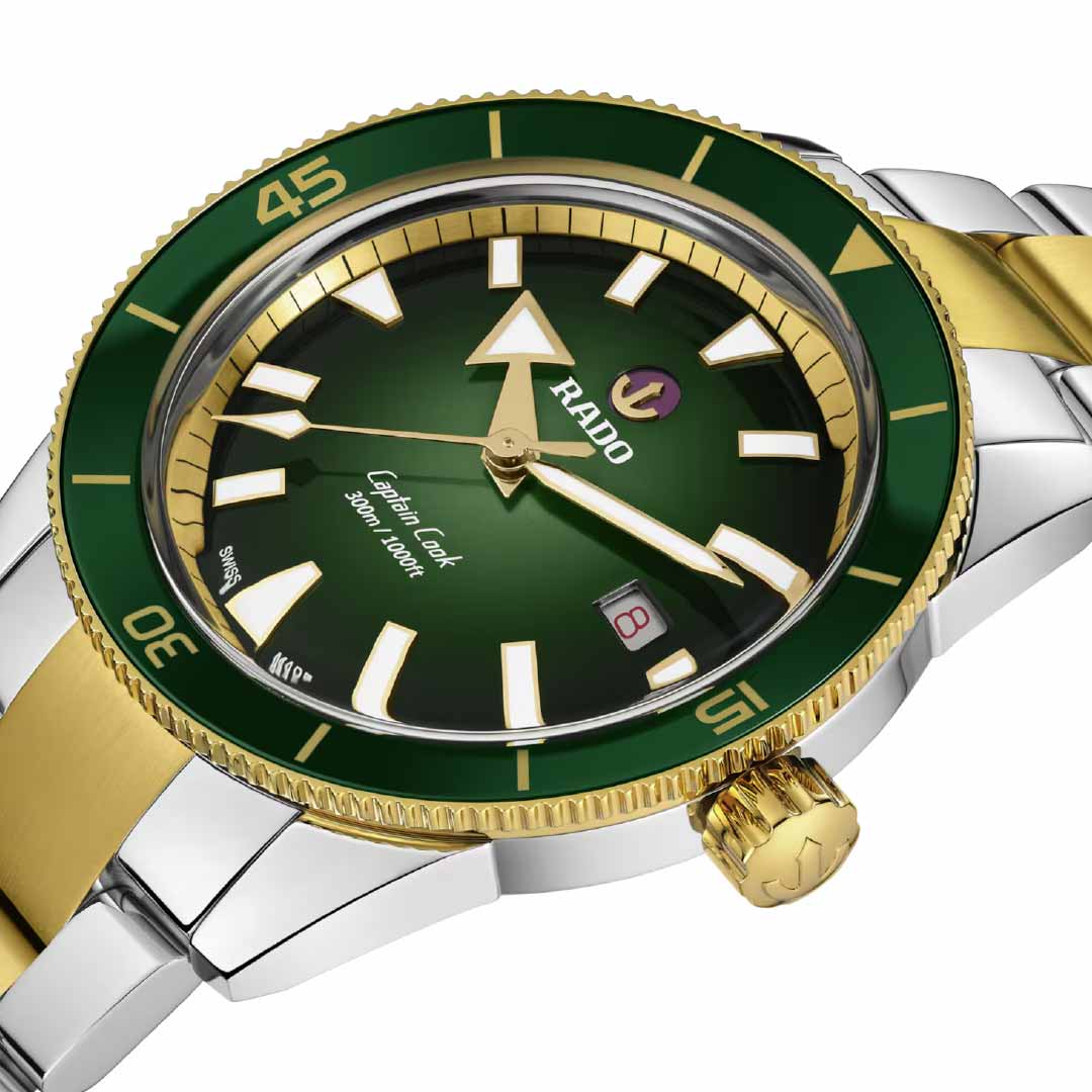 The RADO Captain Cook Automatic 42mm Watch showcases a detailed view highlighting its green and gold bezel, complemented by a green dial with white hour markers. It houses the sophisticated Rado R763 automatic movement. The silver and gold stainless-steel bracelet enhances the visible date window at 3 o'clock, with "Rado" prominently featured on the dial.