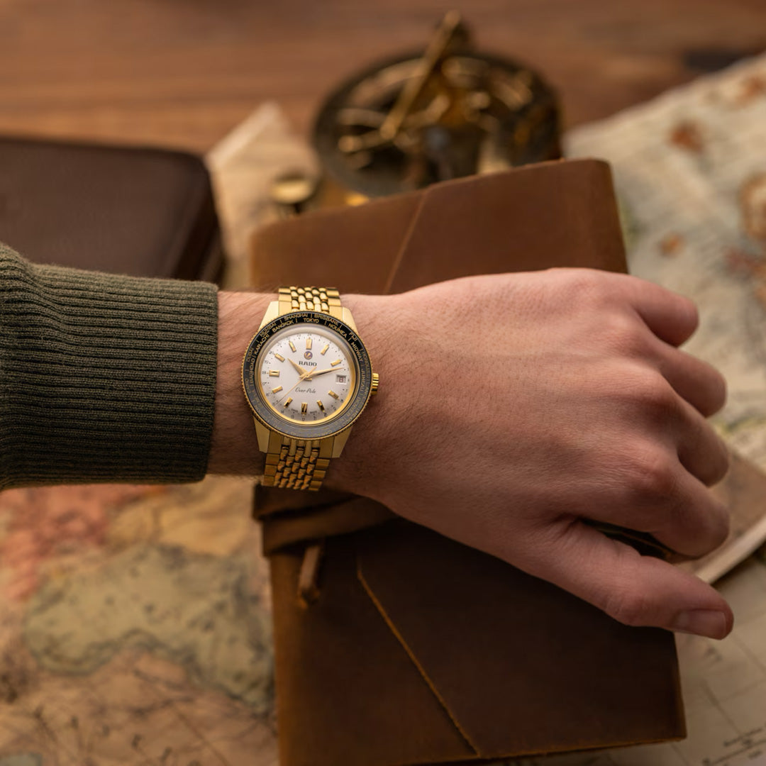 Wearing a green sweater and gold wristwatch, a person rests their hand on a brown leather journal, with a vintage map and instrument in the background—showcasing the RADO Captain Cook Over-Pole Limited Edition Handwinding 39mm Watch.