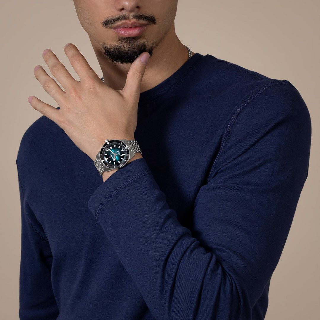 A man in a navy blue long-sleeve shirt touches his face with his left hand, showcasing the RADO Captain Cook Automatic 39mm Watch by RADO, featuring a dark detailed dial. The background is a neutral beige color.