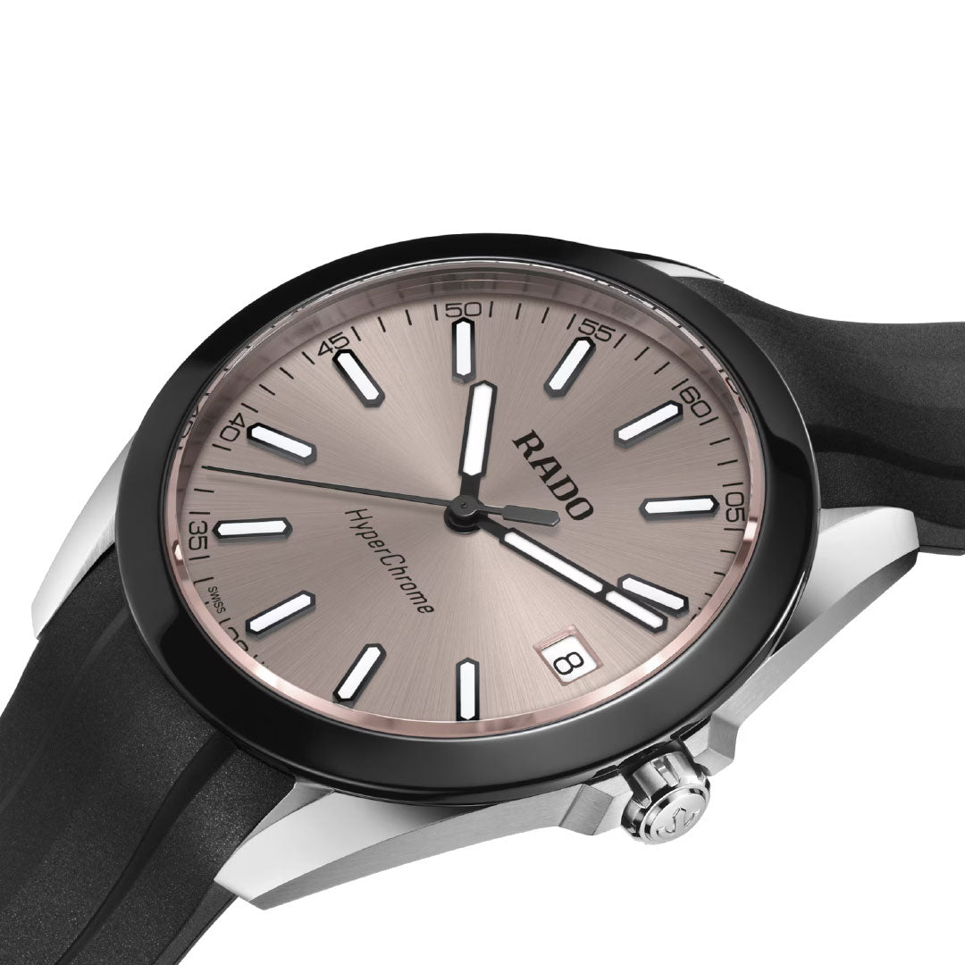 A close-up of the RADO HyperChrome Quartz 41.5mm Watch, featuring a rose gold dial, white hour markers, high-tech ceramic bezel, black rubber strap, and date display at the 3 o’clock position.