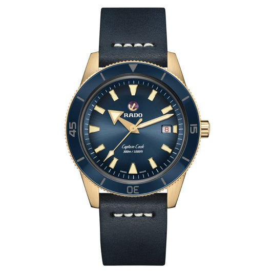 The RADO Captain Cook Automatic Bronze 42mm Watch showcases a captivating gold-tone case paired with a blue face. It is equipped with prominent luminous markers and includes a date window at the 3 o'clock position, complemented by a black leather strap. The bezel features minute markings, with RADO's iconic logo elegantly situated below the 12 o'clock marker.