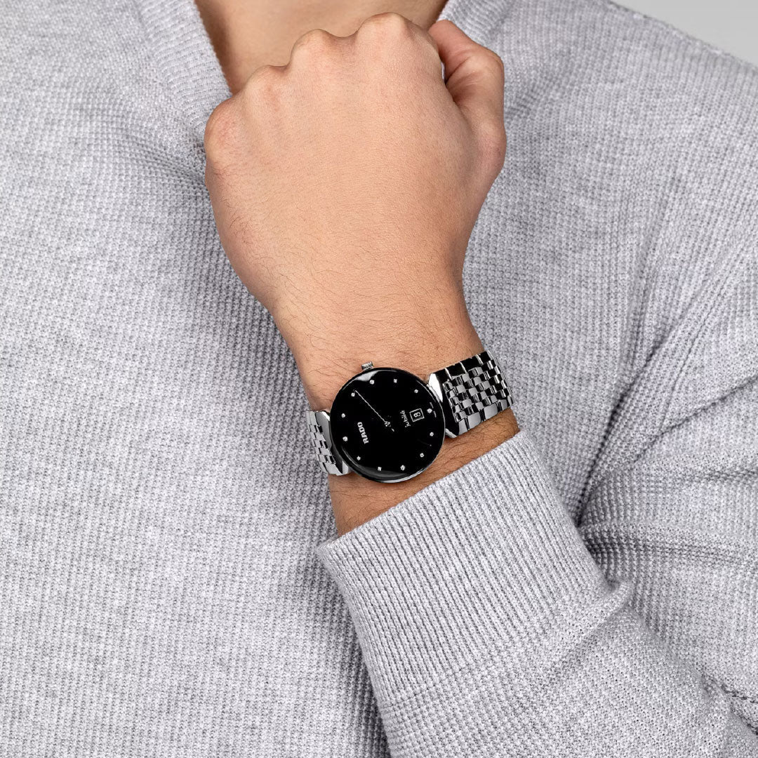 A person in a light gray sweater bends their arm, showing a RADO Florence Diamonds Quartz 38mm Watch with a black face and metal band, displaying the time and date.