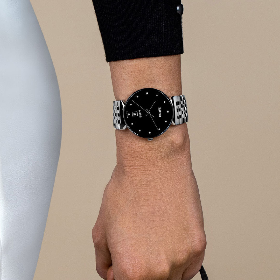A close-up of a person's wrist wearing the RADO Florence Diamonds Quartz 38mm Watch, featuring a black face and dot indices, set against a beige background. The person is dressed in black and white clothing.