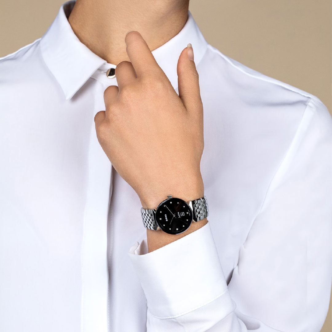 Wearing a crisp white button-up, a person touches their neck, showcasing the RADO Florence Diamonds Quartz 30mm Watch with a sleek black dial on their left wrist. The background is plain and neutral.