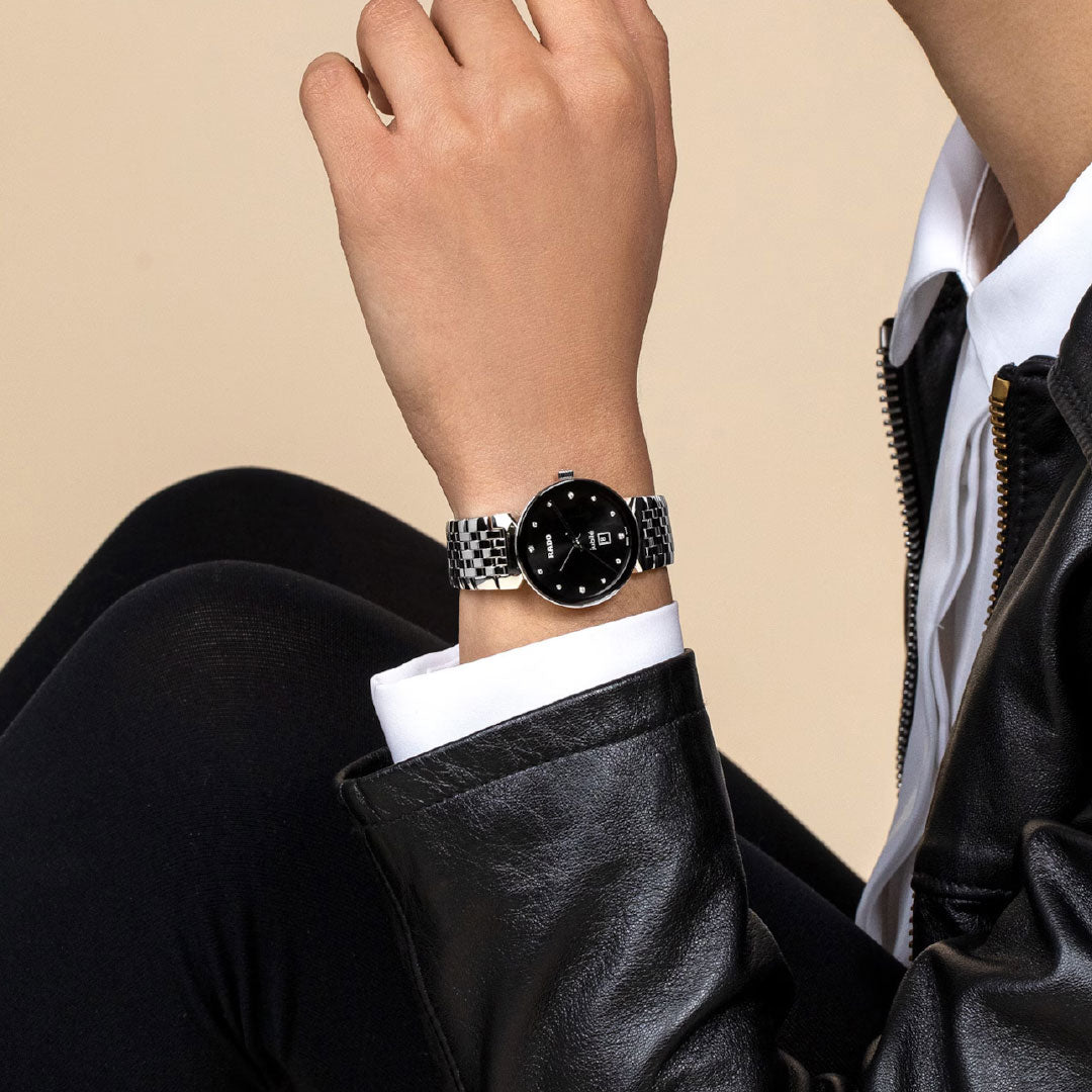 A person in a black leather jacket and white shirt poses against a beige background, highlighting the RADO Florence Diamonds Quartz 30mm Watch with a sleek black dial.