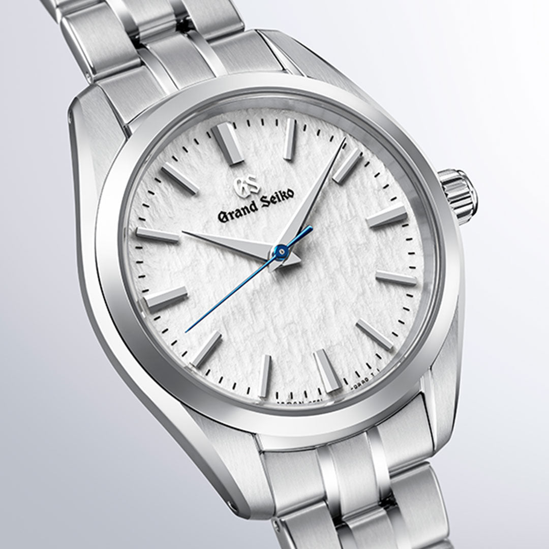 A close-up of the Grand Seiko Heritage Collection 9F51 Quartz 33mm Watch showcases its textured white dial inspired by the Hotaka mountain range, complemented by silver hour markers and a blue second hand. The watch features a stainless steel case and bracelet and is powered by Grand Seiko’s high-precision Caliber 9F51 movement.