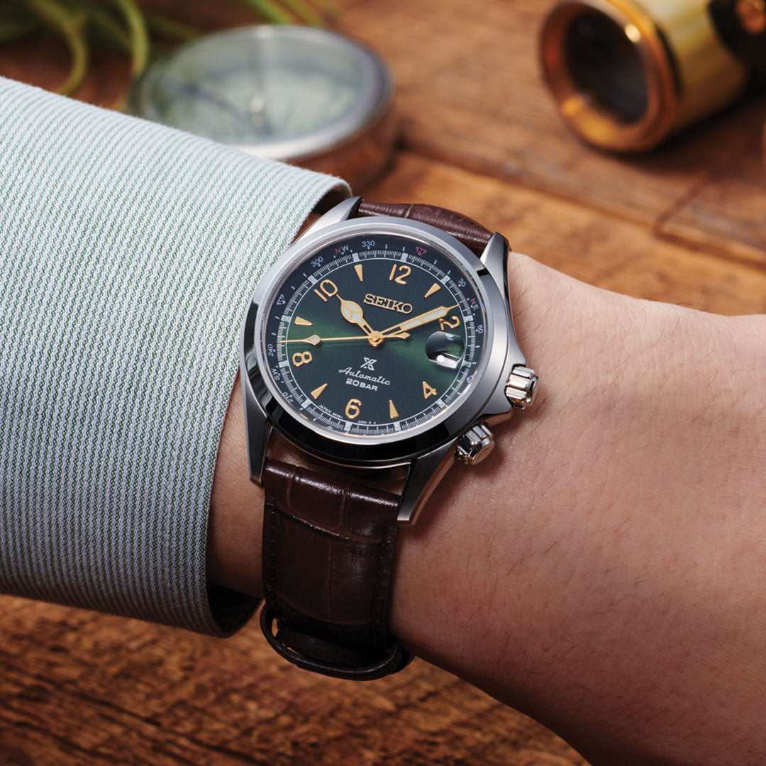 A wrist adorned with a Seiko Prospex Land Automatic With Manual Winding 39.5mm Watch showcases its green dial, gold hands, and brown leather strap. The background features a wooden table with a compass and another partially visible object.