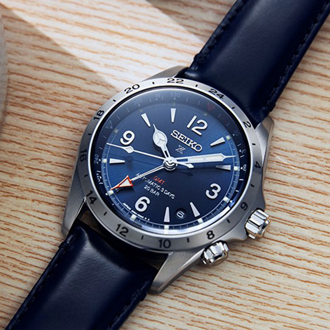 The Seiko Prospex Land Automatic With Manual Winding 39.5mm watch showcases a blue dial paired with a silver bezel, featuring a mechanical GMT function highlighted by a red-tipped hand and powered by the Caliber 6R54 movement. It is elegantly complemented by a blue leather strap and displayed on a light wooden surface.