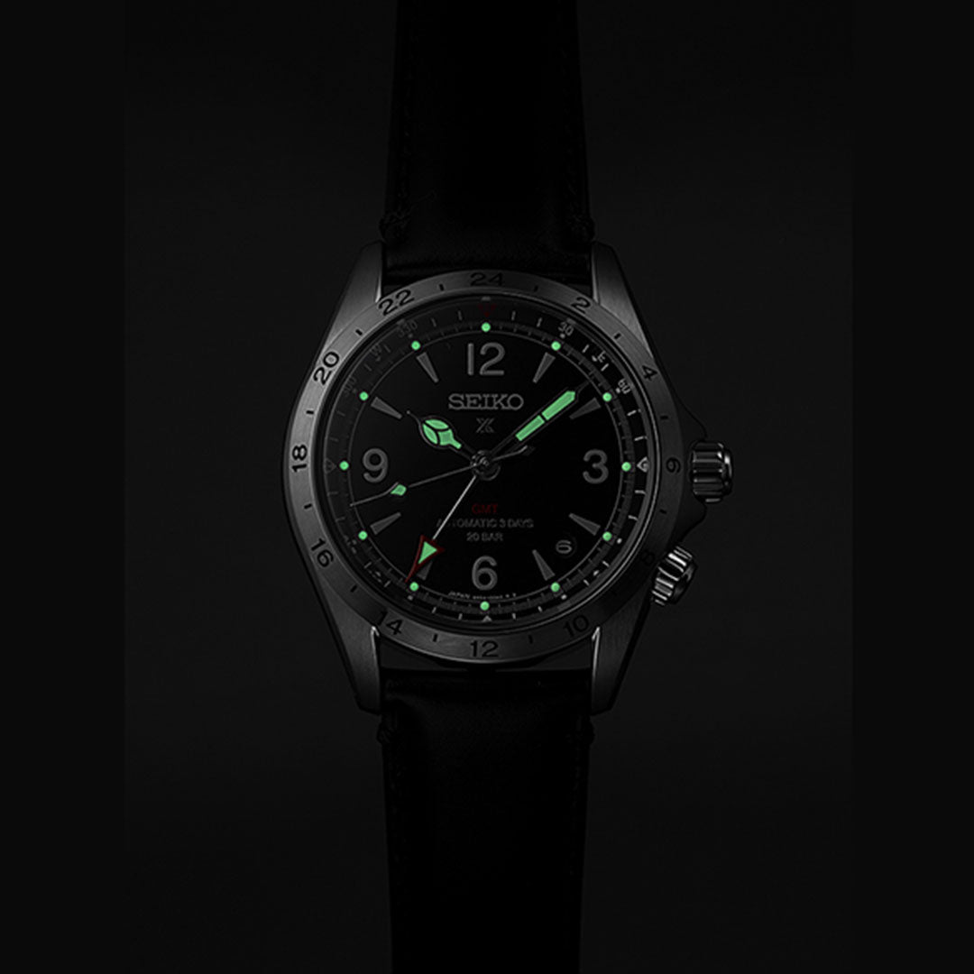 Close-up of a Seiko Prospex Land Automatic With Manual Winding 39.5mm watch, showcasing a dark face and glowing green hands. The watch, which includes the Caliber 6R54 movement, displays the Seiko logo and is equipped with a black strap. The dark background highlights its luminescent elements.