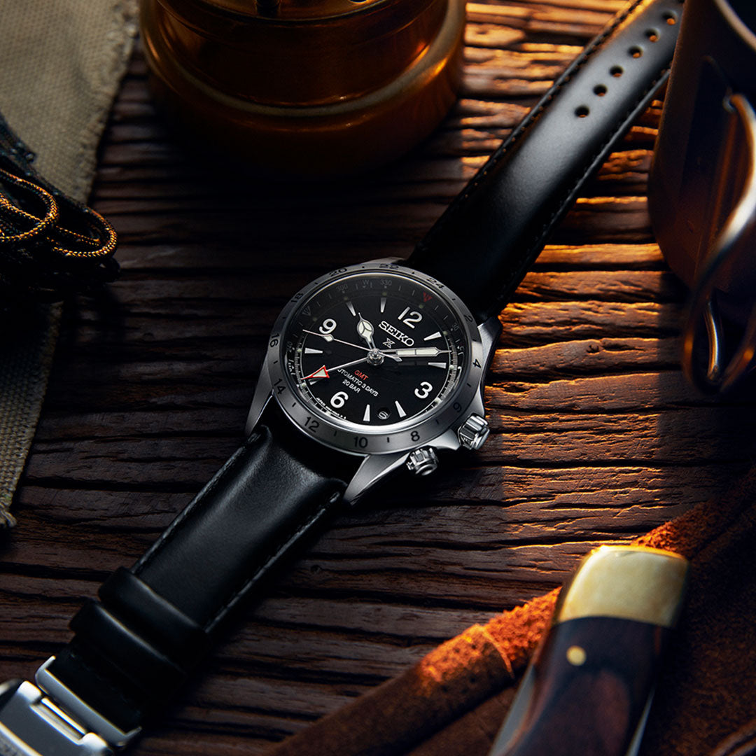 A Seiko Prospex Land Automatic with Manual Winding 39.5mm watch, featuring a sleek black leather strap and the Mechanical GMT function powered by the Caliber 6R54 movement, rests on a wooden surface. Nearby, a knife, a piece of fabric, and a mug create an adventurous rustic atmosphere.