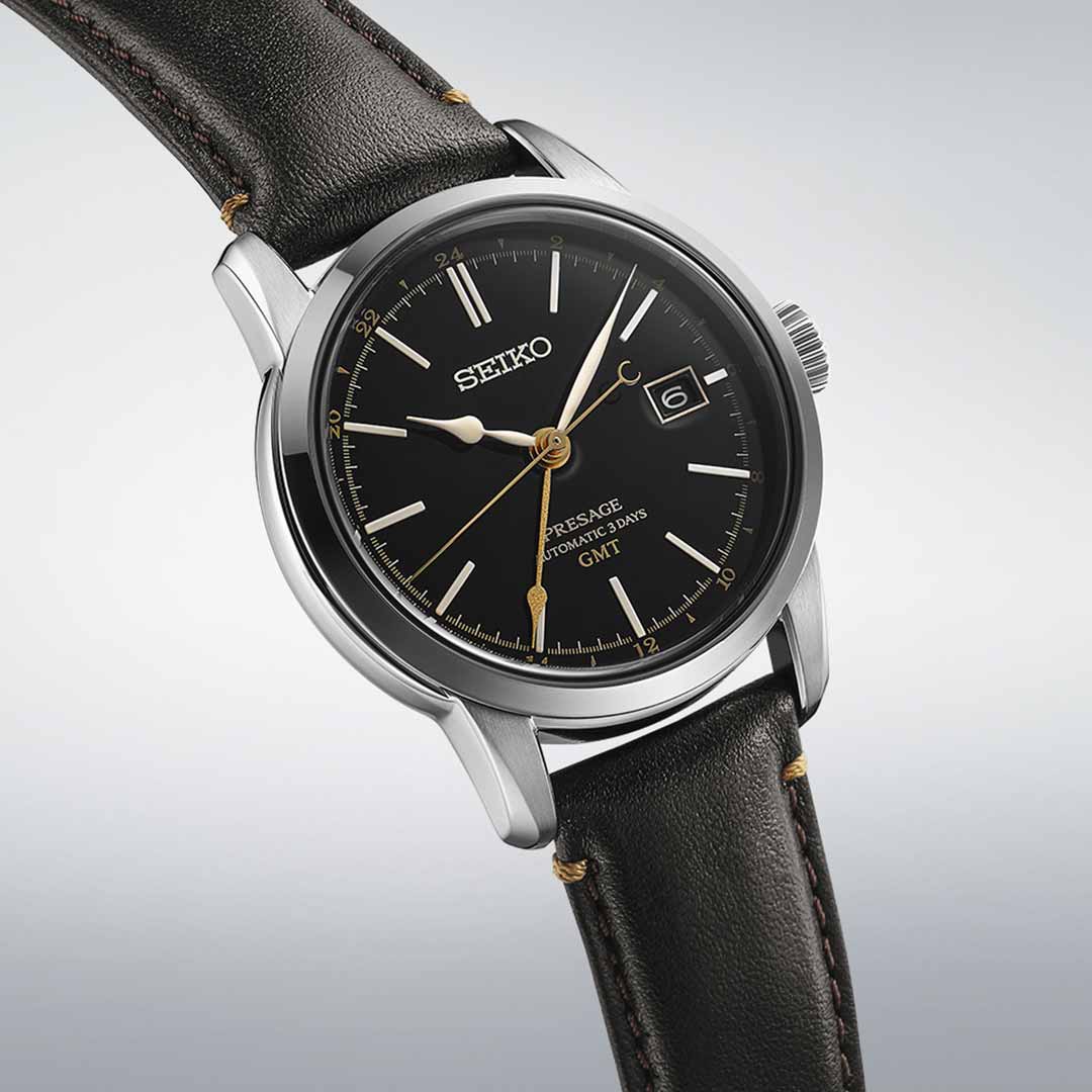 The Seiko Presage Craftsmanship Series Automatic with Manual Winding GMT 40.2mm Watch showcases a black urushi lacquer dial complemented by gold-tone hands and markers, along with a date window at the 3 o'clock position. This exquisite timepiece features a brown leather strap and silver-tone case, elegantly set against a light gradient background.
