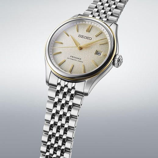 The Seiko Presage Classic Series Automatic 40.2mm watch features a silver and gold-tone bezel, textured light dial, date window at 3 o’clock, and a stainless steel bracelet, showcased against a plain gradient background.