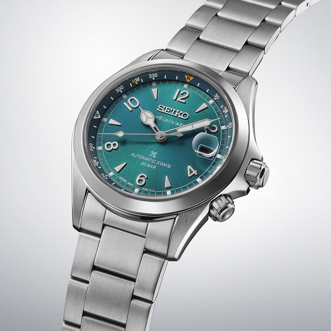A Seiko Prospex Land Automatic With Manual Winding 39.5mm watch by Seiko, showcasing a stainless steel case, teal dial with silver hour markers, date window, and metal bracelet, is elegantly displayed on a light background.