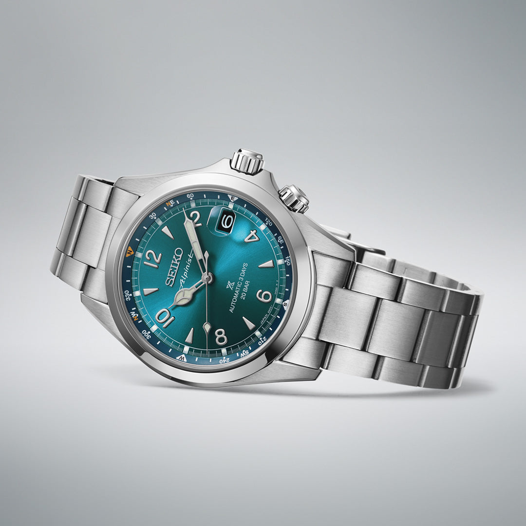 A Seiko Prospex Land Automatic With Manual Winding 39.5mm Watch by Seiko, featuring a stainless steel case, teal dial, silver hour markers, and a date display, is displayed on a plain light grey background—blending precision with timeless style.