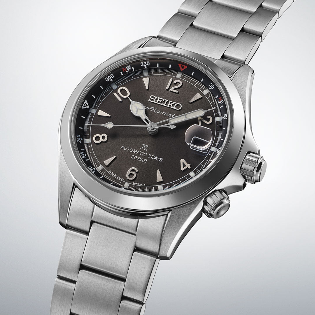 The Seiko Prospex Land Alpinist Automatic With Manual Winding 39.5mm Watch features a stainless steel case, grey dial with date window, silver markers and hands, brushed metal bracelet, and two crowns, set against a light gradient background.
