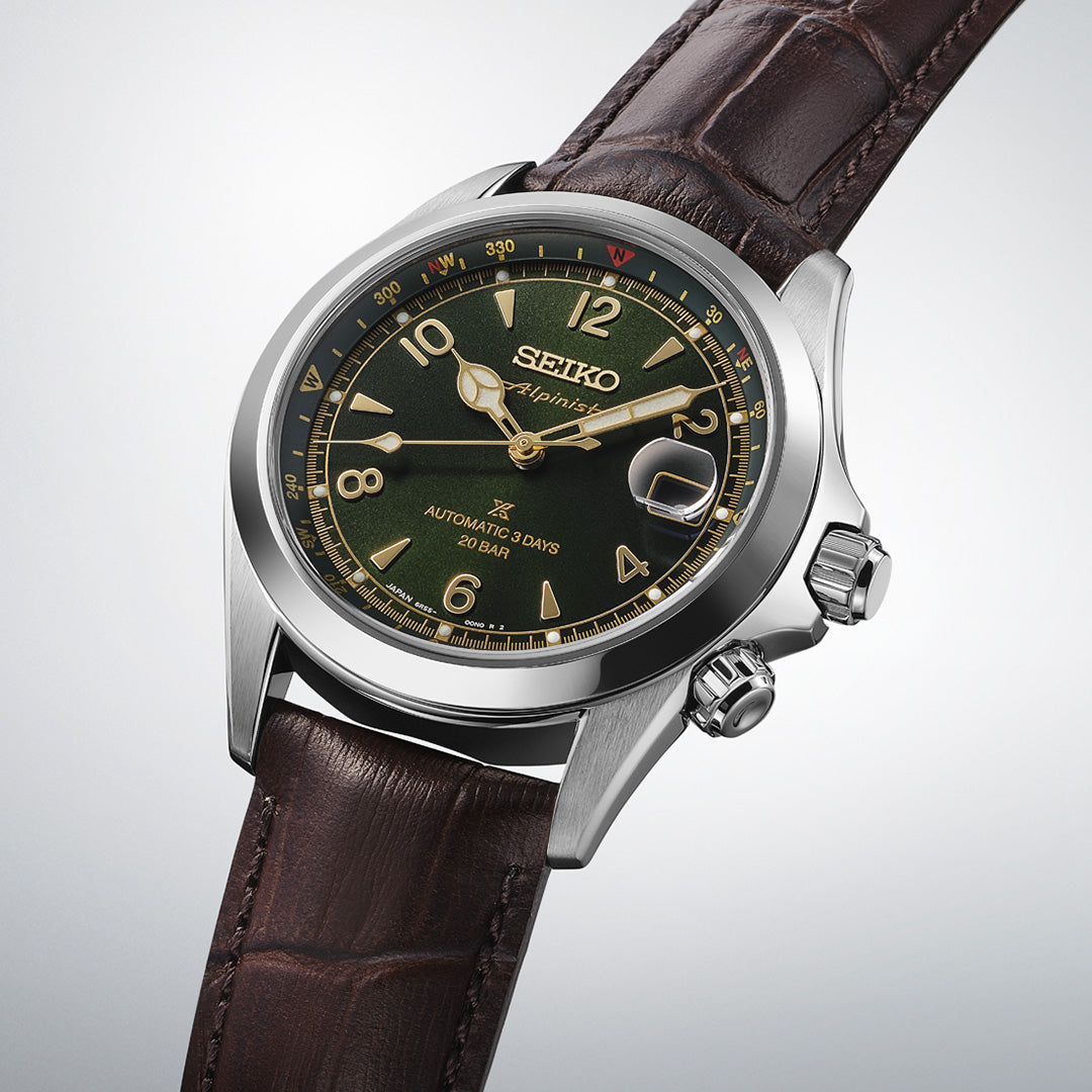 A Seiko Prospex Land Automatic With Manual Winding 39.5mm Watch by Seiko, featuring a dark green dial with gold accents and a brown leather strap, is displayed at an angle against a light background.